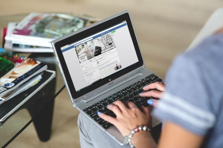 A person using a laptop, browsing a social media profile. Nearby, there are magazines on a glass table. The setting appears to be a casual indoor environment, with the person wearing a gray t-shirt and a bracelet.