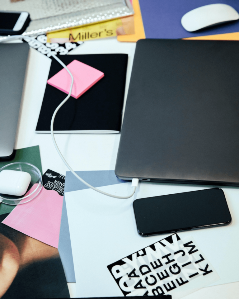 A cluttered desk with a laptop, smartphone, charging cable, assorted stationery, a mouse, colorful paper sheets, earphones in a case, and a book titled 