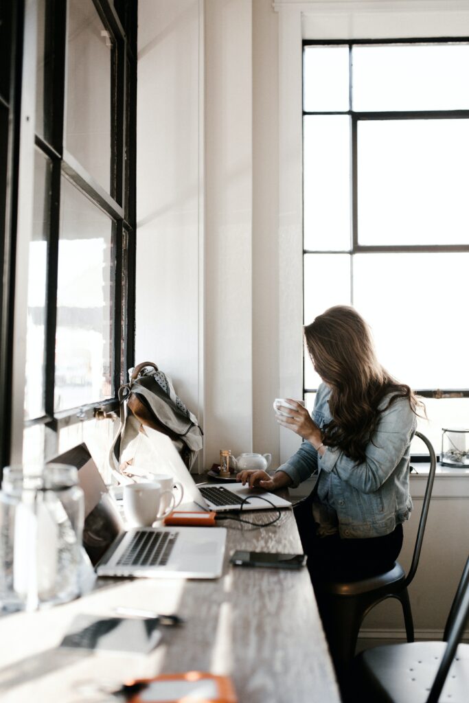 A person with long hair sits at a window-side table in a bright room, holding a cup and looking at a laptop. A backpack, water bottle, and other items are on the table. Sunlight streams through the large window.