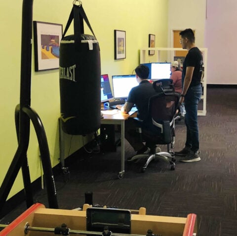 A room with a punching bag and foosball table in the foreground. Three people are seated at desks working on computer screens while one person stands behind them. The walls are light green with framed photos hanging on them.
