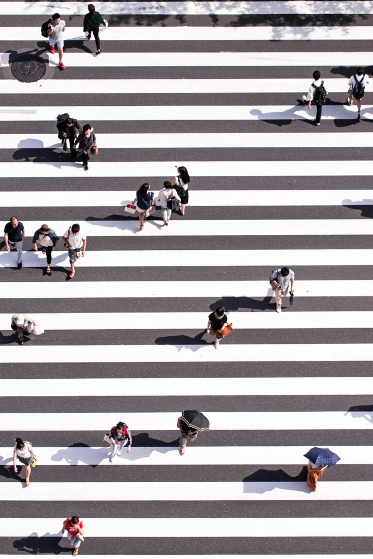 Aerial view of a busy pedestrian crossing with people walking in various directions. The crosswalk consists of wide, alternating black and white stripes. Some individuals carry umbrellas, while others hold bags. Shadows stretch across the striped path.