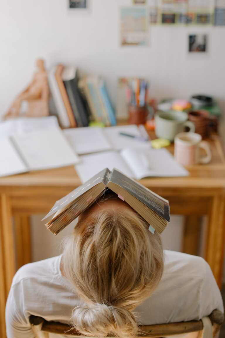 A person with light hair is sitting at a desk, resting their head backward. An open book is placed on their face. The desk is cluttered with books, papers, stationery, and a mug. The background shows wall decorations.
