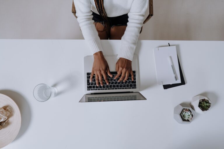 A person wearing a white sweater types on a laptop at a white desk. Nearby are a glass of water, two small potted plants, and a stack of papers with a pen. The person sits on a light chair, with braided hair visible. Top-down view.