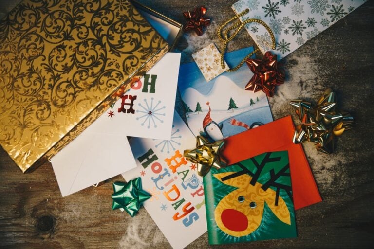 A collection of holiday-themed greeting cards with various designs, including snowflakes, a reindeer, and a snowman. They are surrounded by colorful bows, a golden envelope, and decorative ribbon on a wooden surface dusted with snow.