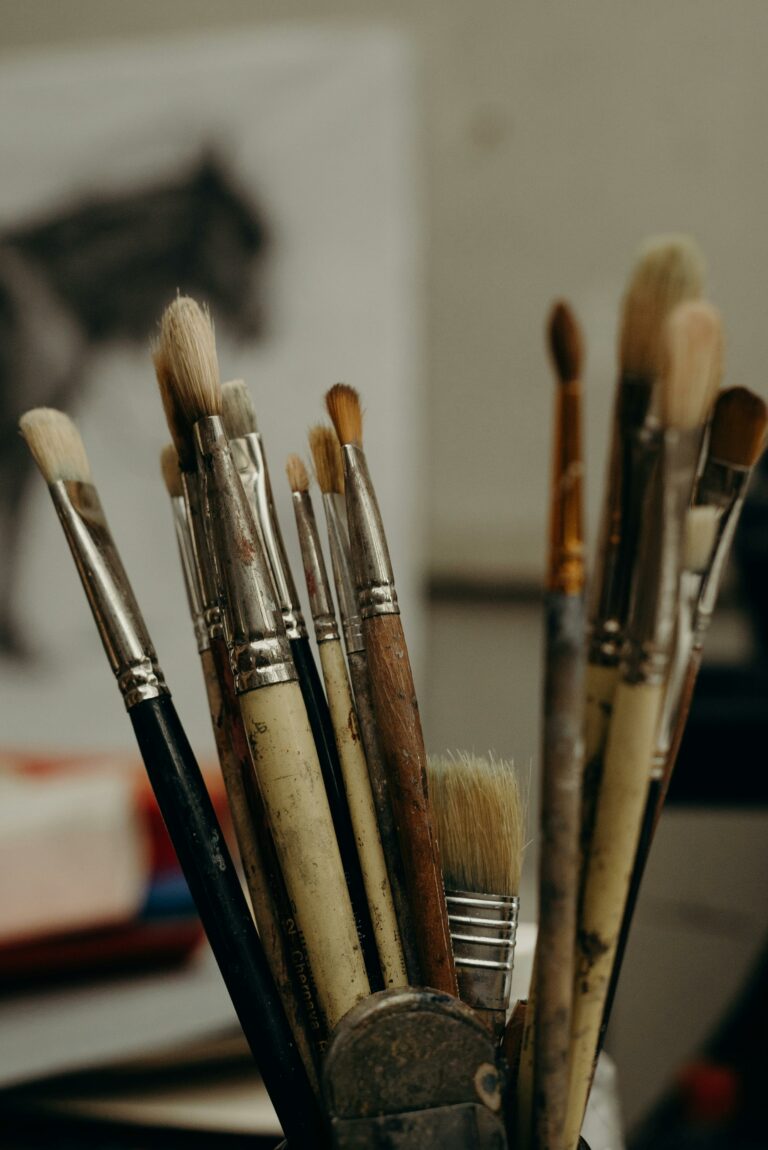 A collection of paintbrushes of varying sizes and styles stands upright in a metal holder. The bristles show signs of use, and a blurred charcoal sketch of a horse is visible in the background.