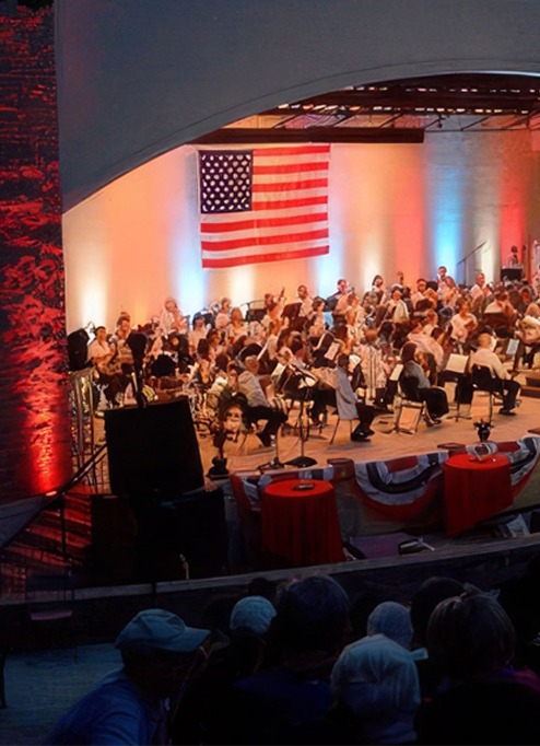 An orchestra performs on an outdoor stage adorned with red, white, and blue lighting. A large American flag hangs as a backdrop. The audience watches from the foreground, and the venue features a curved roof structure. Various decorations are visible.