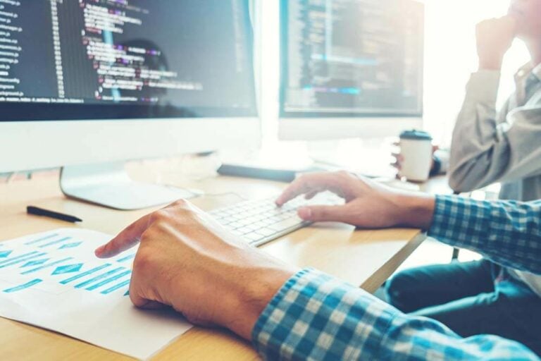 Two people are seated in front of computer monitors filled with lines of code