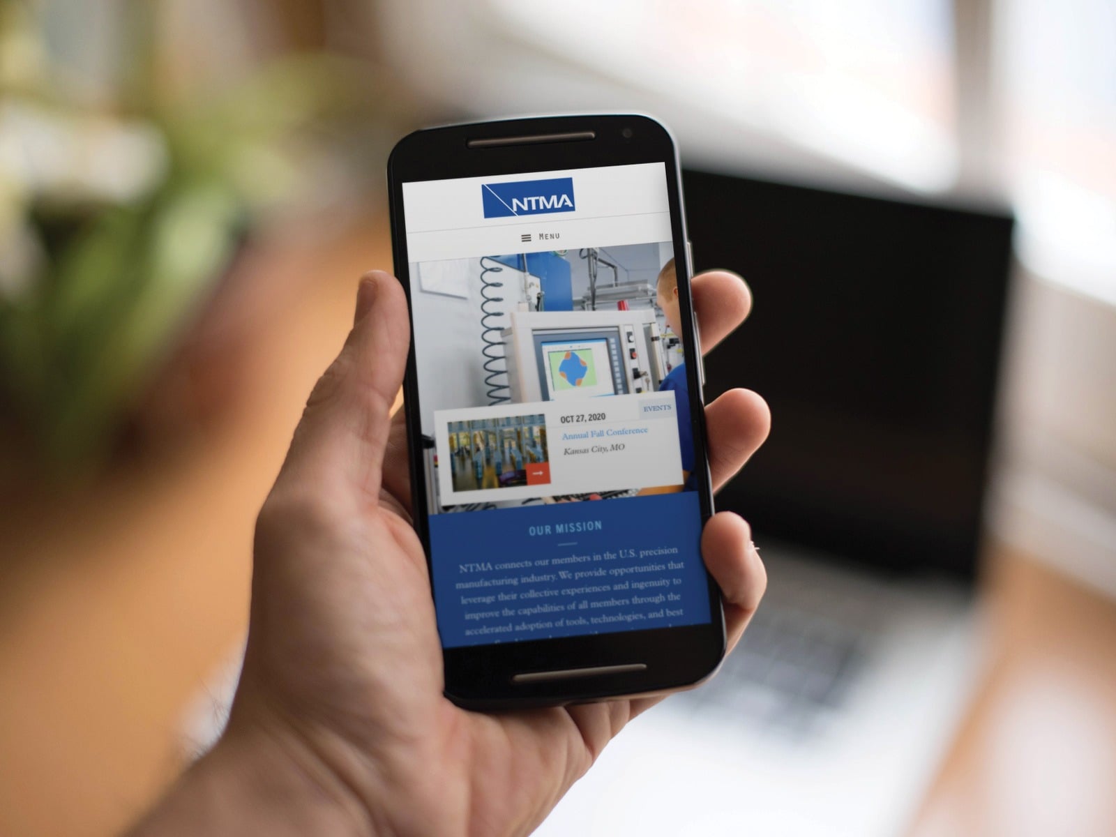 A person holds a smartphone displaying a website homepage for NTMA, showcasing a conference event listing. In the background, a blurred laptop is visible on a table. The setting appears to be indoors near a window.