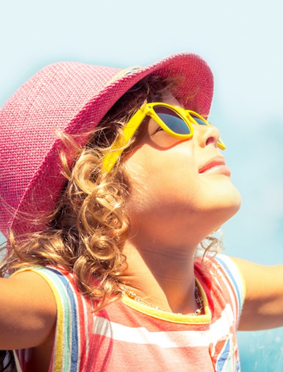 A child faces the sun, wearing yellow sunglasses and a pink hat.