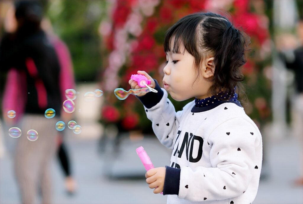 A young child with dark hair is blowing soap bubbles outdoors. Wearing a white sweatshirt with small hearts, the child is intently focused on the bubbles. In the background, there are blurred people and colorful flowers.