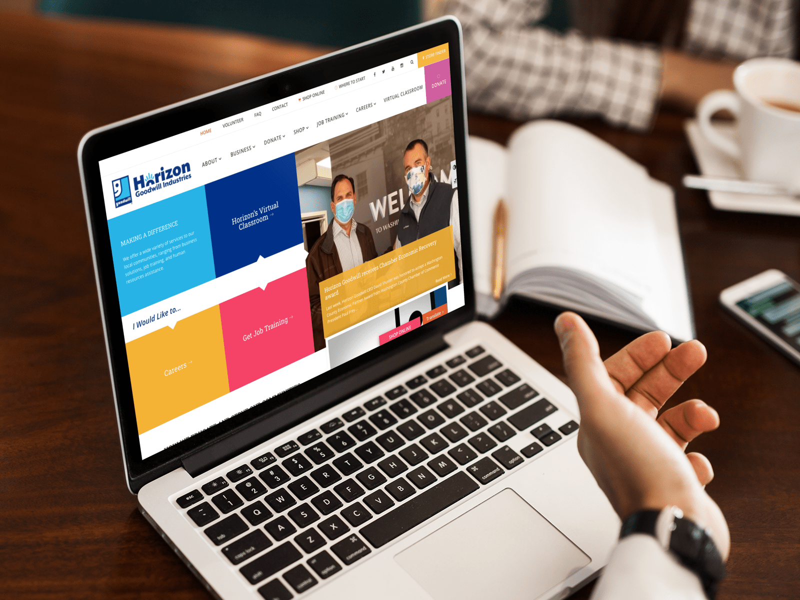 A person gestures at a laptop displaying a colorful website with text and images. Two men in masks appear in a photo on the screen. An open notebook and a cup are on the table.