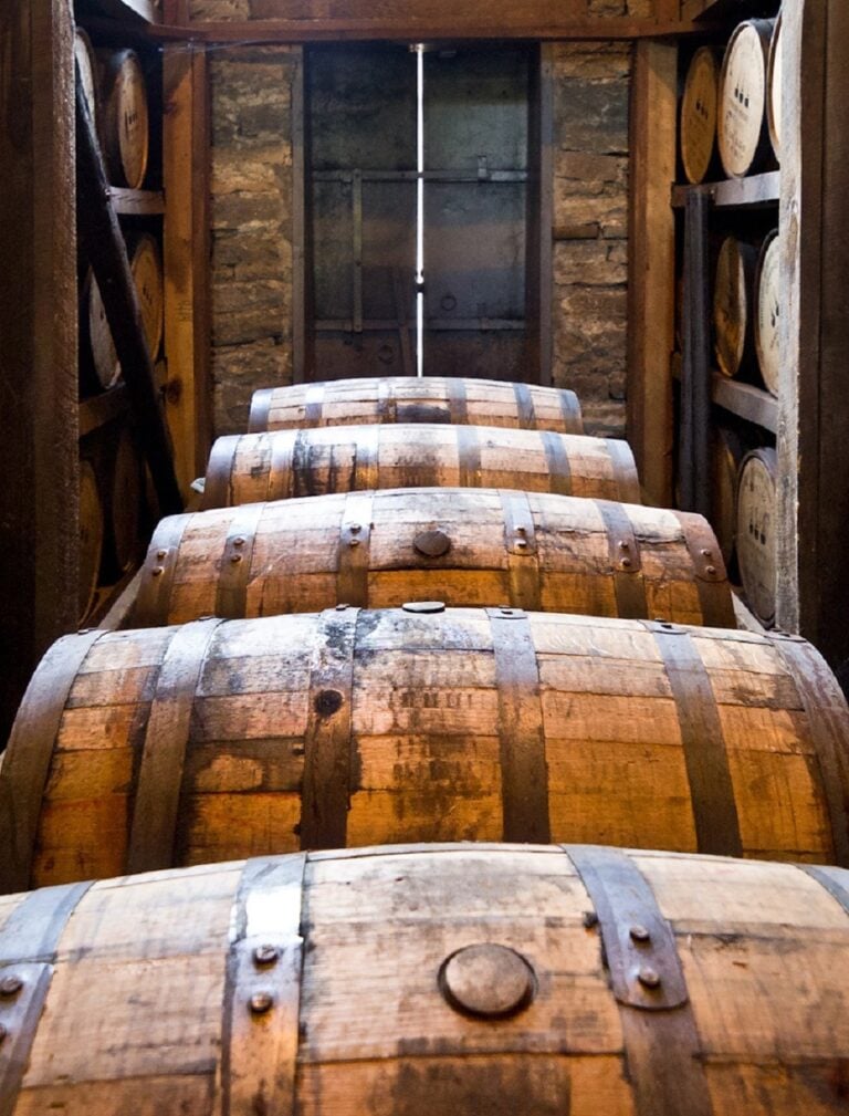 A row of large wooden barrels are stacked in a dimly lit storage room. The barrels are weathered, with metal bands securing them. Stone walls and more barrels in the background suggest a rustic setting, likely a cellar or distillery.
