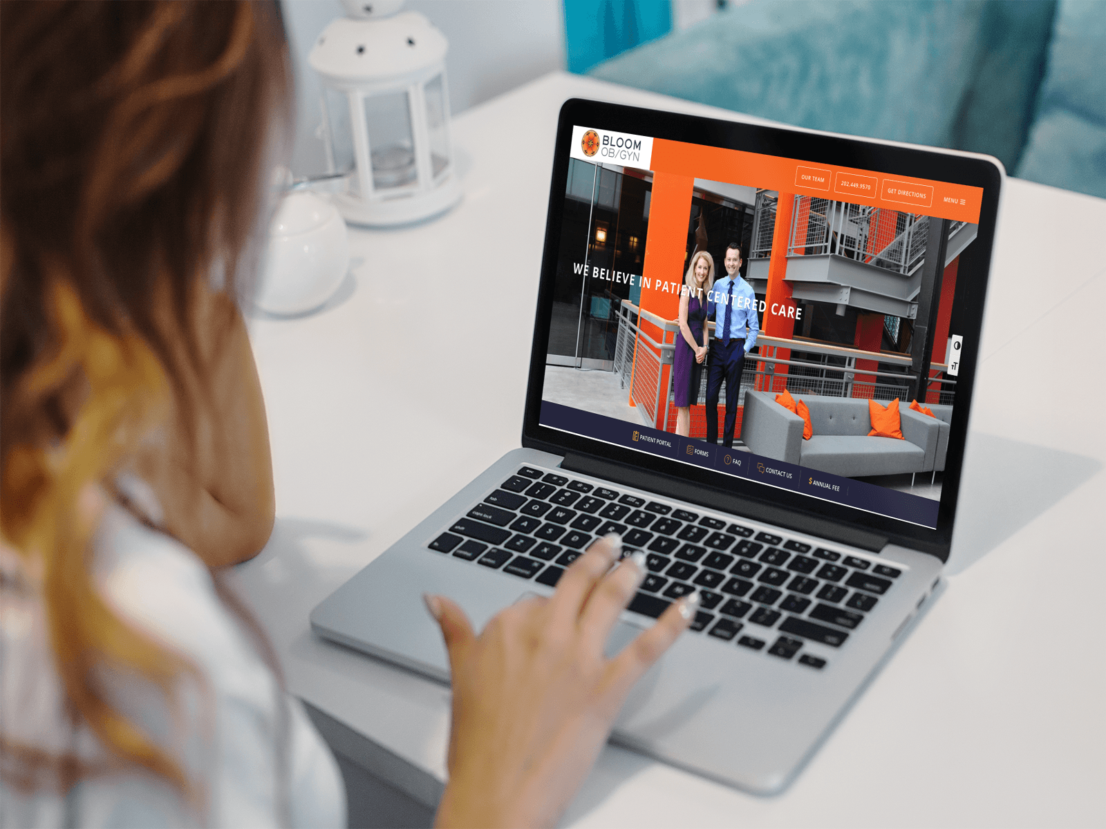 A person with reddish-brown hair sitting at a table using a silver laptop. The screen displays a website with a photo of two people standing in front of a modern building. There is text on the screen related to patient care.