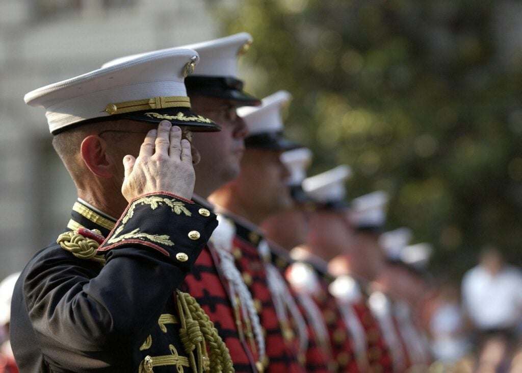 Several uniformed military personnel stand in a row, saluting. The focus is on one individual in the foreground wearing a white cap and a dark dress uniform with gold trim. Others in similar attire are lined up behind, slightly out of focus.