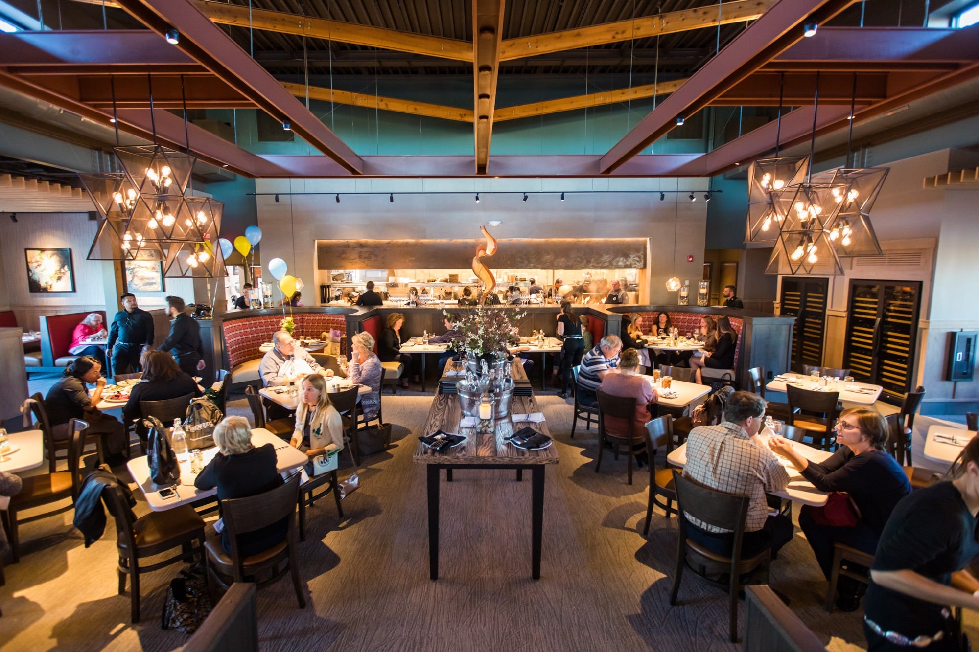 A busy, modern restaurant with diners seated at tables, warm lighting from chandeliers, open kitchen in the background, and staff serving food. The atmosphere is lively and welcoming.