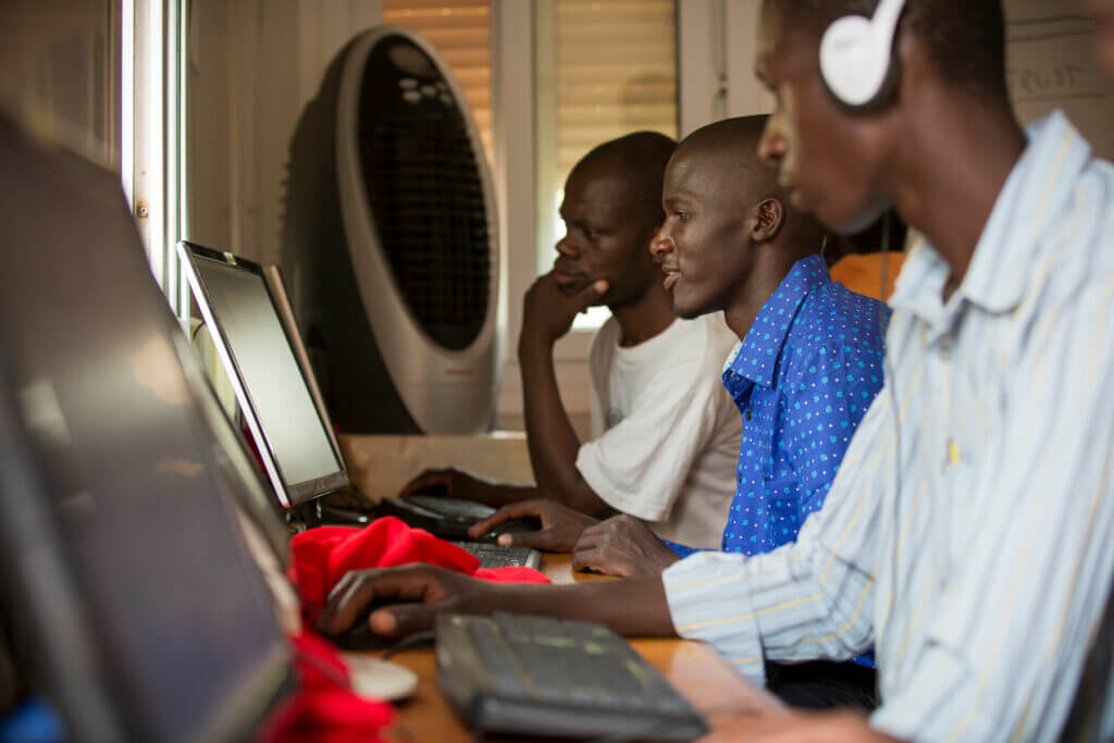 Three men sitting side by side work on desktop computers. One wears headphones and a striped shirt, another wears a blue shirt, and the third wears white. The setting appears to be a computer lab or office.