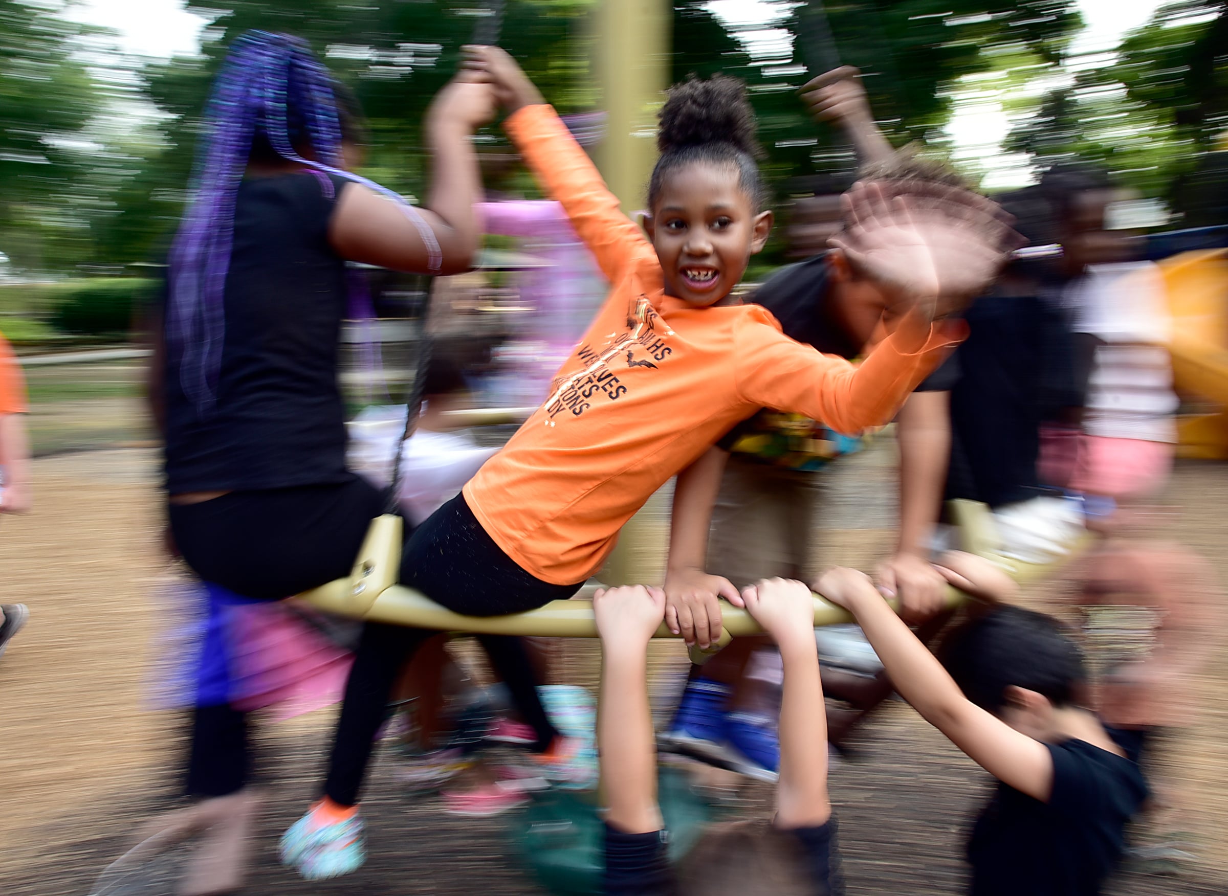 A group of children play energetically on playground equipment. One girl in an orange shirt smiles and waves at the camera while spinning, surrounded by other blurred, moving children.
