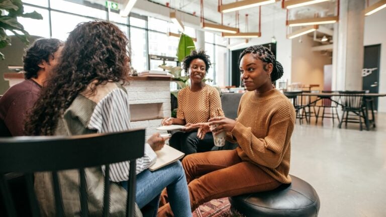 Four people sit together in a modern, well-lit office space, engaged in conversation. Two women face each other, one gesturing while holding a glass, and notebooks are open as they discuss or collaborate.