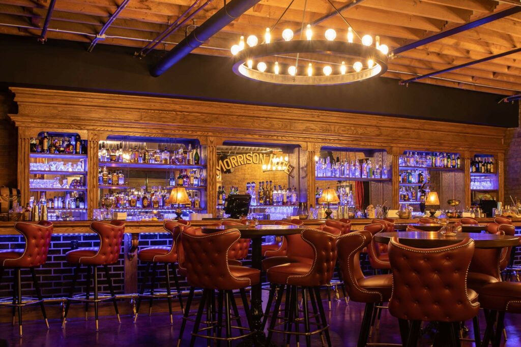 A stylish bar with tufted red leather chairs, wooden countertops, shelves stocked with bottles, soft yellow lighting, and a large circular chandelier hanging from a wooden ceiling.