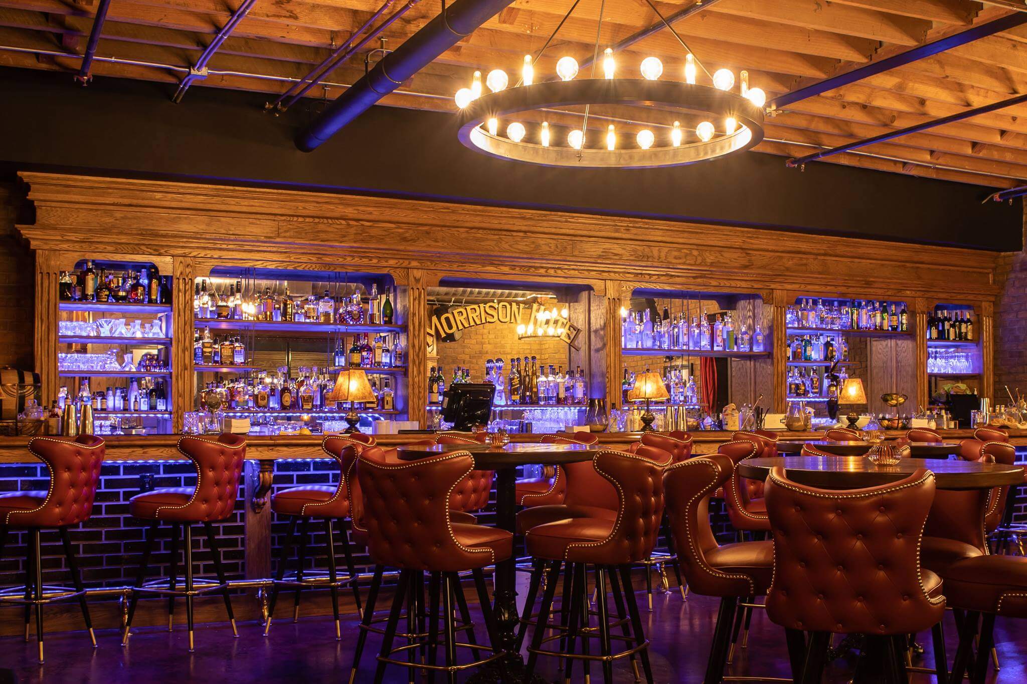 A stylish bar with tufted red leather chairs, wooden countertops, shelves stocked with bottles, soft yellow lighting, and a large circular chandelier hanging from a wooden ceiling.