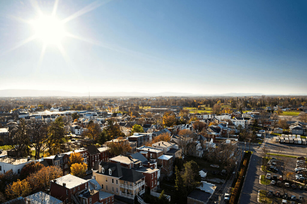 Aerial view of Hagerstown's Bester community, a small neighborhood with houses, trees, and streets under a bright, sunny sky. The sun shines intensely, casting clear shadows, and distant hills are visible on the horizon.