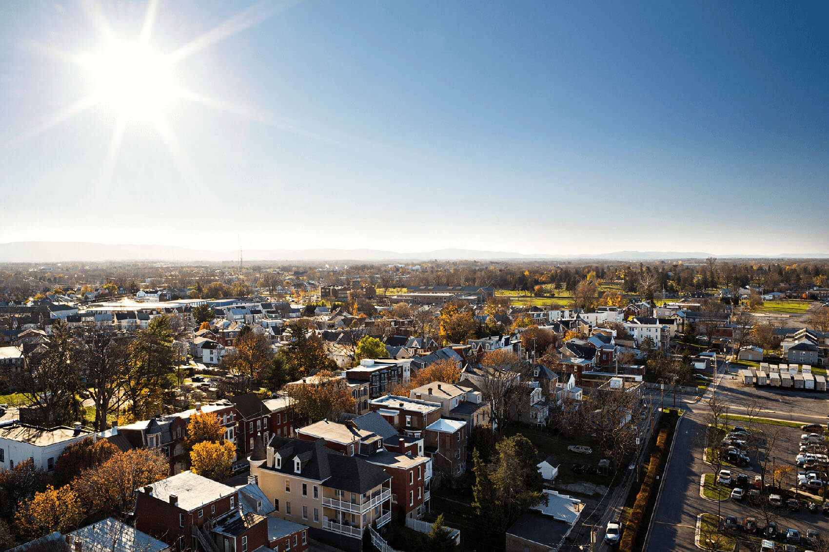 Aerial view of Hagerstown's Bester community, a small neighborhood with houses, trees, and streets under a bright, sunny sky. The sun shines intensely, casting clear shadows, and distant hills are visible on the horizon.