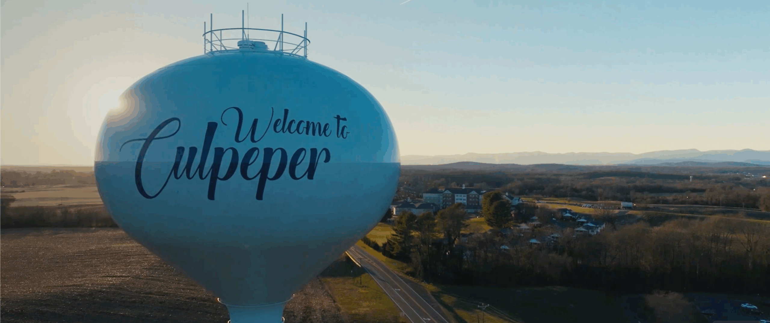 A large water tower with the words “Welcome to Culpeper” stands beside a rural highway, overlooking open fields and distant mountains during sunrise or sunset.