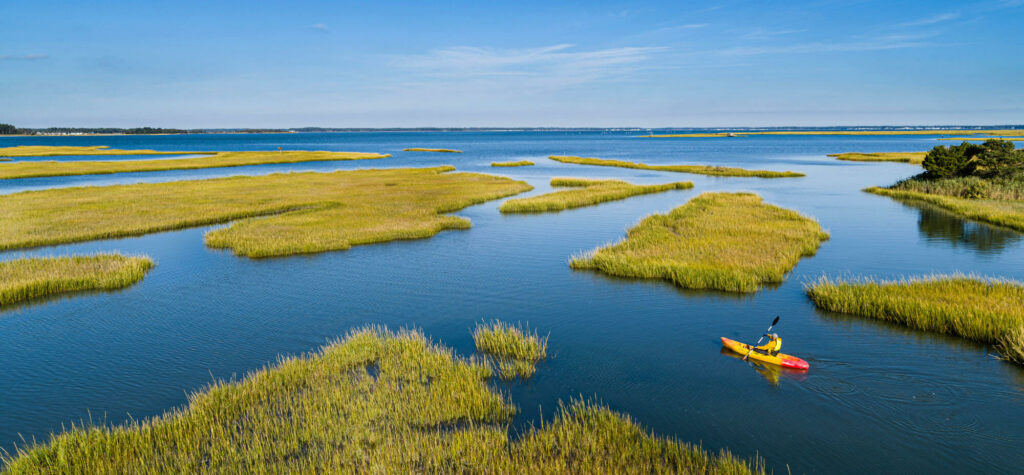 A person in a yellow kayak paddles through calm blue water among grassy marsh islands under a clear sky.