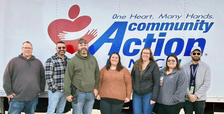 Seven smiling people stand in front of a large sign that reads 