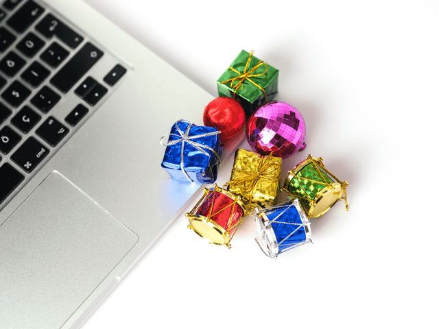 A laptop keyboard beside small, colorful holiday Christmas ornaments—including gift boxes, drums, a red ball, and a pink disco ball—on a white background.