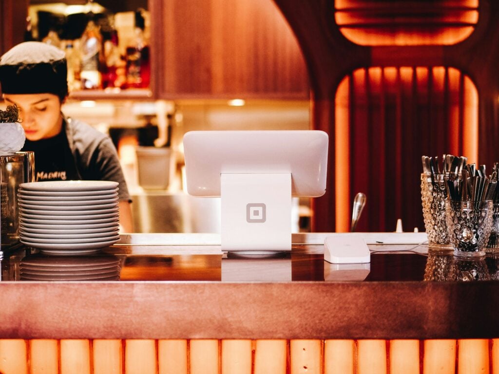 A cash register with a tablet screen sits on a wooden counter, surrounded by stacked plates and utensils. In the warmly lit restaurant, holiday decorations add a festive touch as a person works in the background.