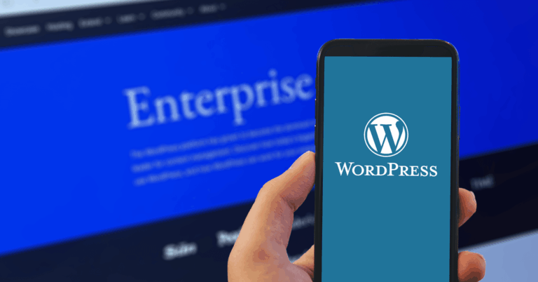 A person holds a smartphone displaying the WordPress logo on its screen, with a blurred computer monitor in the background showing the word 