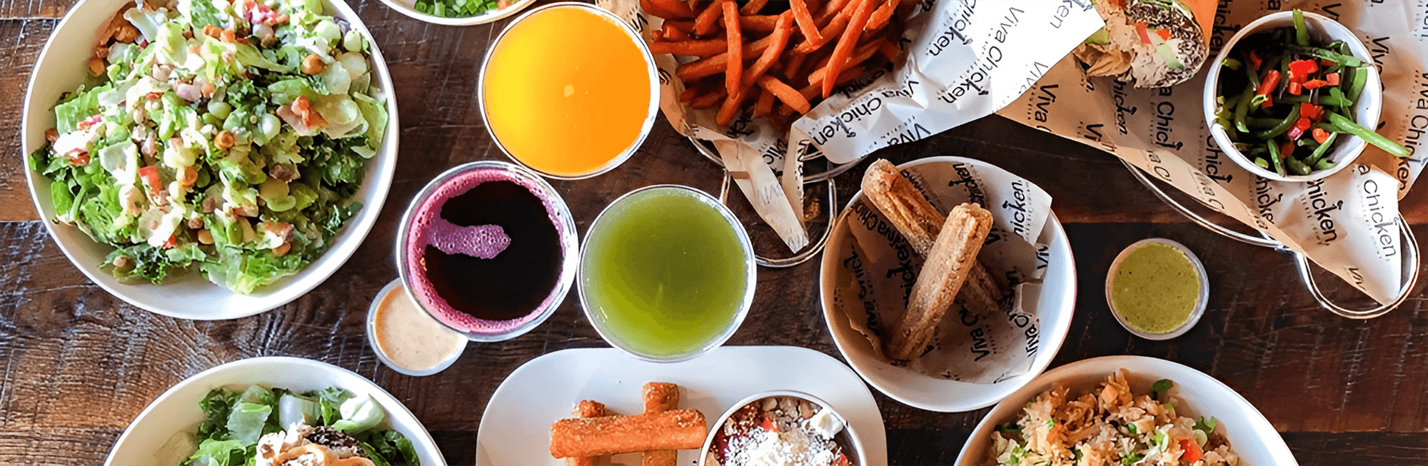 A wooden table topped with a variety of colorful dishes, including salads, fries, bread, dips, and vibrant drinks, showcasing a fresh and diverse meal spread.