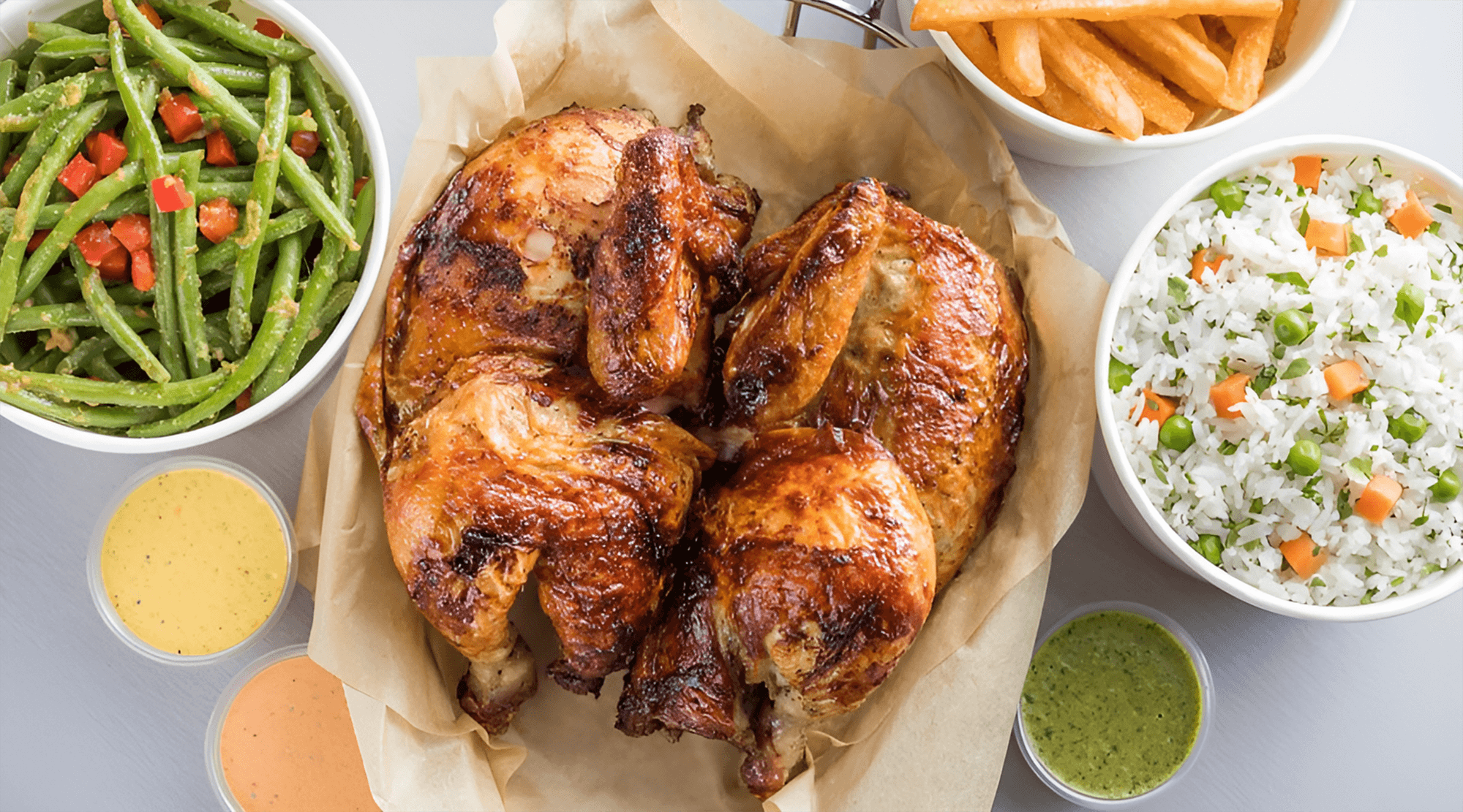 A roasted chicken is served on parchment paper, surrounded by bowls of green beans with diced tomatoes, French fries, rice with vegetables, and four small containers of various sauces.