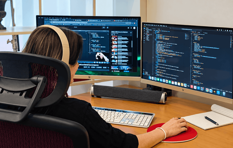A person wearing headphones works at a desk with two computer monitors displaying code and a video, using a wireless keyboard and mouse.